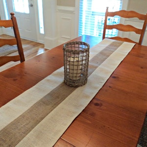 May include: A wooden dining table with a burlap and white striped table runner. A candle is in a wire cage on the table runner.