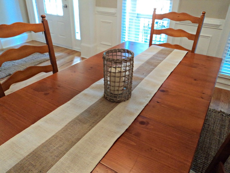 May include: A wooden dining table with a burlap and white striped table runner. A candle is in a wire cage on the table runner.