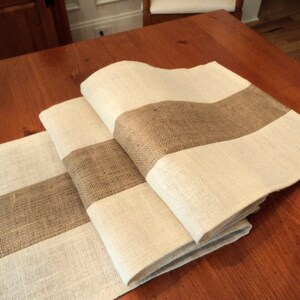 May include: A set of three burlap table runners with white stripes. The runners are folded and arranged on a wooden table.