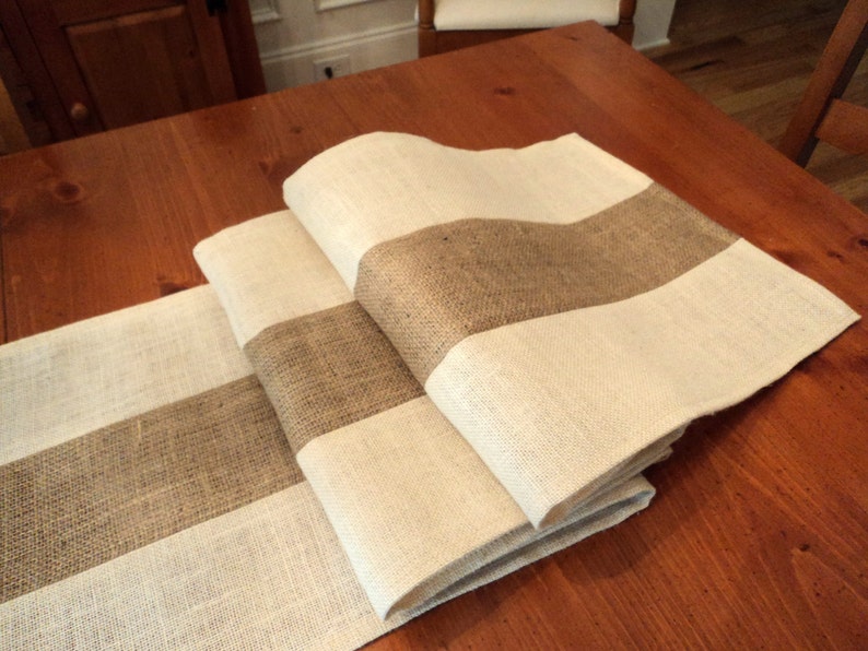 May include: A set of three burlap table runners with white stripes. The runners are folded and arranged on a wooden table.