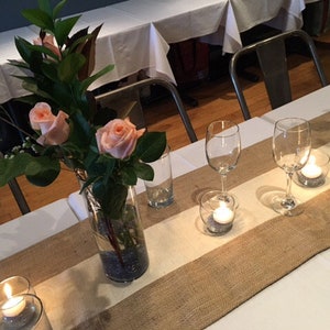 Pode incluir: Uma toalha de mesa branca com um corredor de juta e um vaso de rosas cor-de-rosa. H&aacute; velas e copos na mesa.