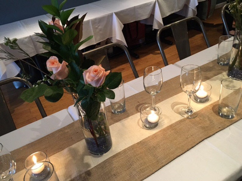 Pode incluir: Uma toalha de mesa branca com um corredor de juta e um vaso de rosas cor-de-rosa. H&aacute; velas e copos na mesa.