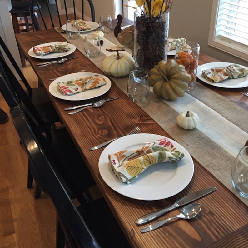 Pode incluir: Uma mesa de madeira r&uacute;stica preparada para um jantar de A&ccedil;&atilde;o de Gra&ccedil;as com pratos brancos, guardanapos com tema de outono e ab&oacute;boras e abobrinhas decorativas.