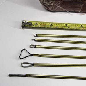 Könnte beinhalten: Eine Sammlung von messingfarbenen Töpferwerkzeugen mit verschiedenen Schlaufen- und Spitzendesigns. Ein gelbes Maßband liegt neben den Werkzeugen und zeigt die Maße in Zentimetern an. Ein verpackter Tonblock befindet sich im Hintergrund.