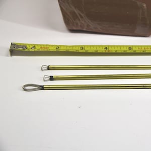 Może przedstawiać: Trzy mosiężne narzędzia pętelkowe do garncarstwa lub rzeźby, z żółtą miarką i brązowym blokiem gliny w tle. Narzędzia mają około 20 cm długości. Narzędzia służą do kształtowania i szczegółowego opracowywania gliny.