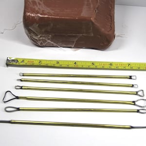 May include: A collection of six brass-colored clay sculpting tools with various tip shapes, including loops and angled edges. A yellow measuring tape shows inches. A wrapped block of brown clay is in the background.