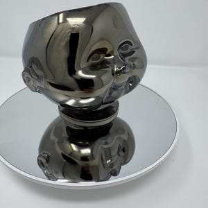 Puede incluir: Una escultura gris metálica de la cabeza de un bebé, con la parte superior ahuecada. La escultura está sobre una superficie redonda y espejada con un borde blanco. La cara del bebé tiene rasgos detallados.