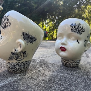 Może przedstawiać: Dwie ceramiczne donice w kształcie głowy lalki. Jedna jest przechylona na bok, druga stoi prosto. Obie są w kolorze ecru z czarnymi wzorami w stylu tatuażu, w tym koronami, ćmą i różami. Usta są pomalowane na czerwono, a podstawa ma geometryczny wzór.