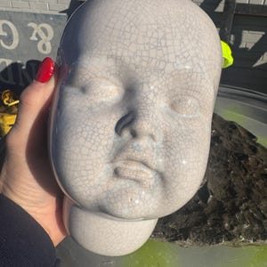 May include: A ceramic baby head sculpture with a crackle glaze finish. The sculpture is off-white with a network of fine gray lines. The head has closed eyes, a small nose, and a slightly downturned mouth. The neck is attached to a rounded base.
