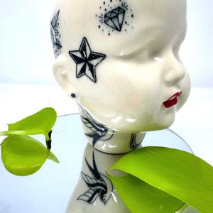May include: A cream-colored ceramic head with black tattoo-style designs, including a star, diamond, and floral elements. The head has red lips and is reflected in a circular mirror, with green leaves in the foreground.