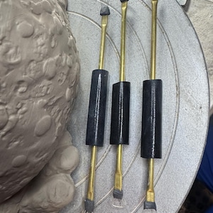 May include: Three pottery tools with brass shafts and black handles are arranged on a pottery wheel. The tools have different shaped tips: a wire brush, a loop, and a rounded edge. A partially visible clay sculpture is in the background.
