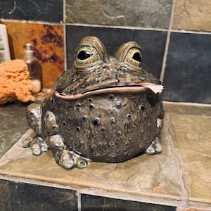 May include: A ceramic frog figurine with a mottled brown and black glaze, featuring prominent bulging eyes with detailed iris patterns and a wide mouth that is slightly open, revealing a small piece of white paper tucked inside the lip.