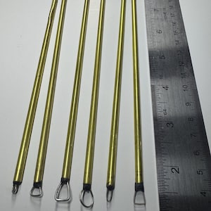 Könnte beinhalten: Sechs messingfarbene Tonmodellierwerkzeuge mit verschiedenen Schlaufen- und Drahtenden. Sie liegen auf einer weißen Oberfläche neben einem Metalllineal. Die Werkzeuge variieren in der Länge, die längsten sind ca. 18 cm lang.