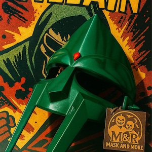 Puede incluir: Una máscara verde angular con una gema roja, sobre un fondo con la palabra "VILLAIN" en amarillo. Una etiqueta marrón con el logotipo de "M&R Mask and More" cuelga de la máscara. El fondo tiene un estilo retro de cómic.