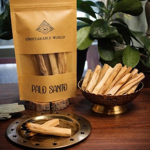 May include: A display of Palo Santo sticks, a natural incense, with a package labeled "Unspeakable World" and "Palo Santo." The sticks are in a decorative bowl and on a small brass plate. The background includes greenery.