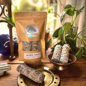 May include: A brown paper pouch labeled Unspeakable World containing white sage, with two sage bundles in a small copper-colored bowl and one sage bundle resting on a round brass plate with star cutouts, all on a wooden surface.