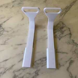 May include: Two identical white L-shaped brackets with a flat base and a vertical arm featuring a rectangular handle cutout at the top. The brackets have a smooth, matte finish and a rigid structure, designed with a wide base for stability.