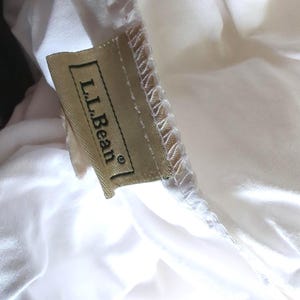May include: Close-up of a white fabric with a sewn seam and a rectangular tan label. The label reads "L.L.Bean" in black text. The fabric is slightly wrinkled and appears to be a light material.