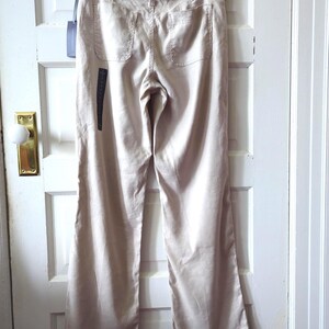 May include: Beige trousers featuring a straight-leg silhouette, a waistband with belt loops, and two back patch pockets. The garment includes a dark brand tag attached to the waistband and a long vertical black product label hanging from the side.
