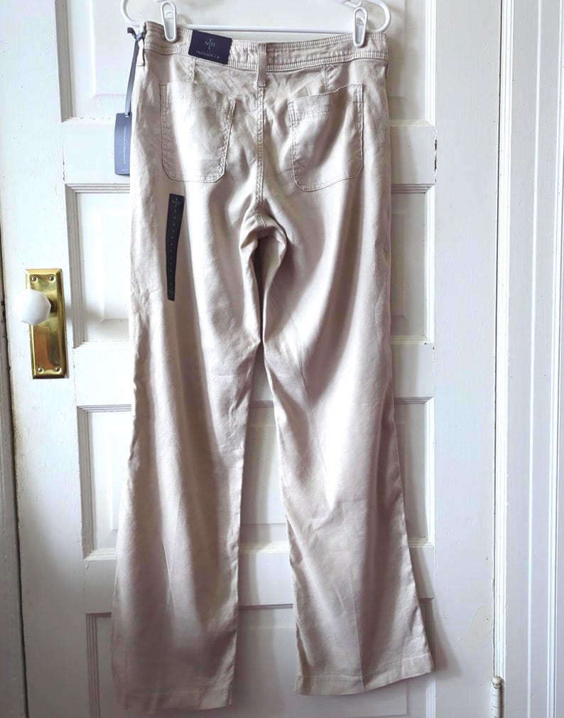 May include: Beige trousers featuring a straight-leg silhouette, a waistband with belt loops, and two back patch pockets. The garment includes a dark brand tag attached to the waistband and a long vertical black product label hanging from the side.