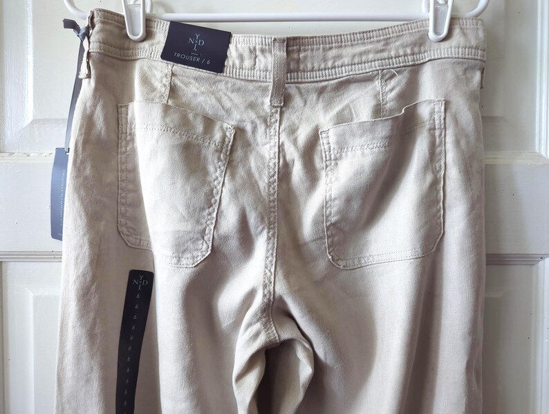 May include: Beige trousers featuring a high waistband with belt loops, two large patch pockets on the back, and a visible brand tag reading YND TROUSER / 8. The garment is displayed on a white hanger with attached retail tags and branding.