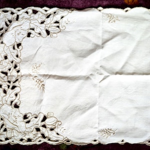 May include: White rectangular table runner with a scalloped edge and embroidered floral and leaf designs. The runner has a detailed cutwork border with a cream-colored thread. The center of the runner is plain white fabric.