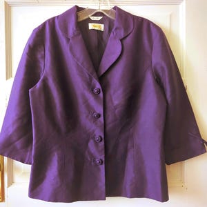 May include: A vibrant purple blazer with three-quarter sleeves and a classic collar. The blazer features a button-down front and tie details at the cuffs. The fabric has a subtle sheen, suggesting a silk or satin material. A tag is visible on the inside of the blazer.