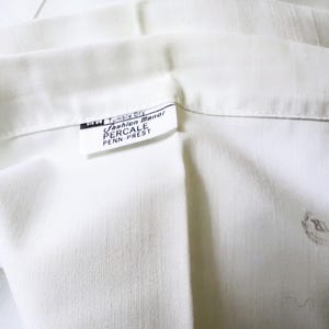 May include: Close-up of white percale fabric with a visible label. The label reads "Fashion Manor PERCALE PENN-PREST" and includes "Tumble Dry" instructions. The fabric is folded, showing texture and subtle variations in the white color.