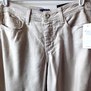 May include: Beige pants featuring a button and zipper fly closure, a waistband with belt loops, two front pockets, and a small coin pocket. A white retail tag is attached to the front, and the pants are suspended from a white plastic hanger.