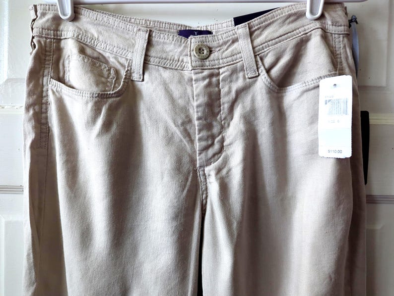 May include: Beige pants featuring a button and zipper fly closure, a waistband with belt loops, two front pockets, and a small coin pocket. A white retail tag is attached to the front, and the pants are suspended from a white plastic hanger.