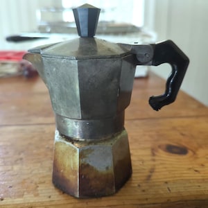 May include: A metallic stovetop espresso maker with an octagonal body, a black plastic handle, and a matching black knob on the lid. The base of the coffee pot shows signs of heat discoloration and wear, with a silver-toned finish throughout.