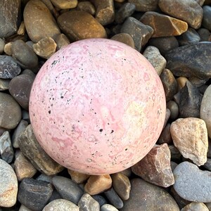 May include: A large, spherical pink stone with dark speckles rests among a bed of various-sized, multi-colored rocks. The smooth, rounded stone is the focal point, contrasting with the rough texture of the surrounding pebbles.