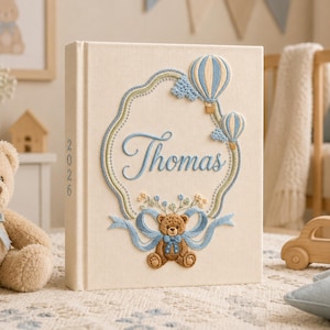 Op de afbeelding: Een crèmekleurig, met stof bekleed fotoalbum met ingewikkeld borduurwerk van de naam Thomas in blauw schrift, omringd door een decoratieve rand met twee heteluchtballonnen, kleine blauwe wolken en een bruine teddybeer met een blauw lint.