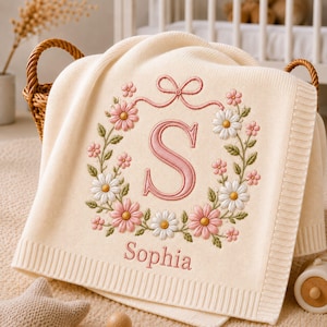 Könnte beinhalten: Eine cremefarbene Strickdecke mit einer Stickerei, die einen großen rosa Buchstaben S in einem Blumenkranz aus rosa und weißen Gänseblümchen zeigt. Über dem Buchstaben befindet sich eine rosa Schleife und darunter der Name Sophia.