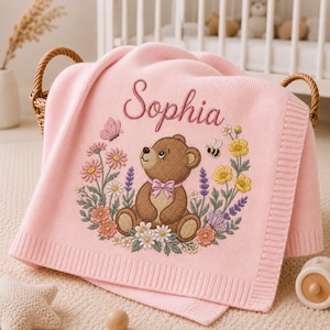 May include: A pink baby blanket embroidered with the name "Sophia". The design includes a teddy bear, flowers, a butterfly, and a bee. The blanket is folded and displayed in a wicker basket. The blanket is made of soft material.