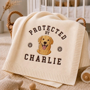 Może przedstawiać: Kremowy koc z dzianiny z wyhaftowanym pośrodku portretem golden retrievera. Nad psem znajduje się napis "PROTECTED BY", a pod nim "CHARLIE", otoczony dwiema małymi ikonami odcisków łap wykonanymi ciemnobrązową nicią.