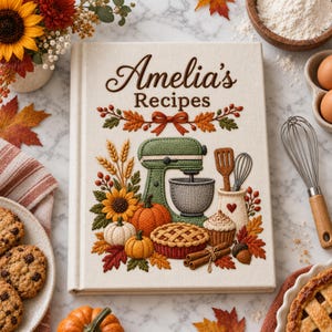 May include: A cream-colored hardcover recipe book titled Amelia's Recipes with embroidered autumn-themed illustrations including a green stand mixer, pumpkins, sunflowers, a pie, cinnamon sticks, and fall leaves on a white marble surface.