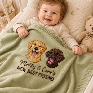 May include: A sage green knitted baby blanket featuring embroidered portraits of a golden retriever and a chocolate labrador. The text "Molly & Coco's NEW BEST FRIEND" is stitched in brown thread below the dog faces on the soft fabric.