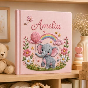 Może przedstawiać: Jasnoróżowy kwadratowy album na zdjęcia z haftowanymi detalami, w tym imieniem Amelia zapisanym kursywą, szarym słoniem, różowym balonem, kolorową tęczą, małymi żółtymi gwiazdkami, motylem oraz różnymi różowymi i białymi kwiatami.