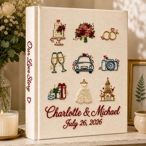 Puede incluir: Un álbum de fotos de tela color crema con iconos de boda bordados: pastel, ramo, anillos, copas de champán, coche, cámara, regalos, vestido e iglesia. El lomo dice Our Love Story, con Charlotte & Michael y la fecha July 26, 2026.