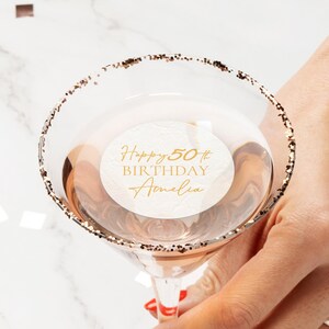 May include: A martini glass with a gold glitter rim, filled with a light-coloured liquid. A white circular sticker inside the glass reads "Happy 50th Birthday Amelia" in gold script. The glass is held by a person.