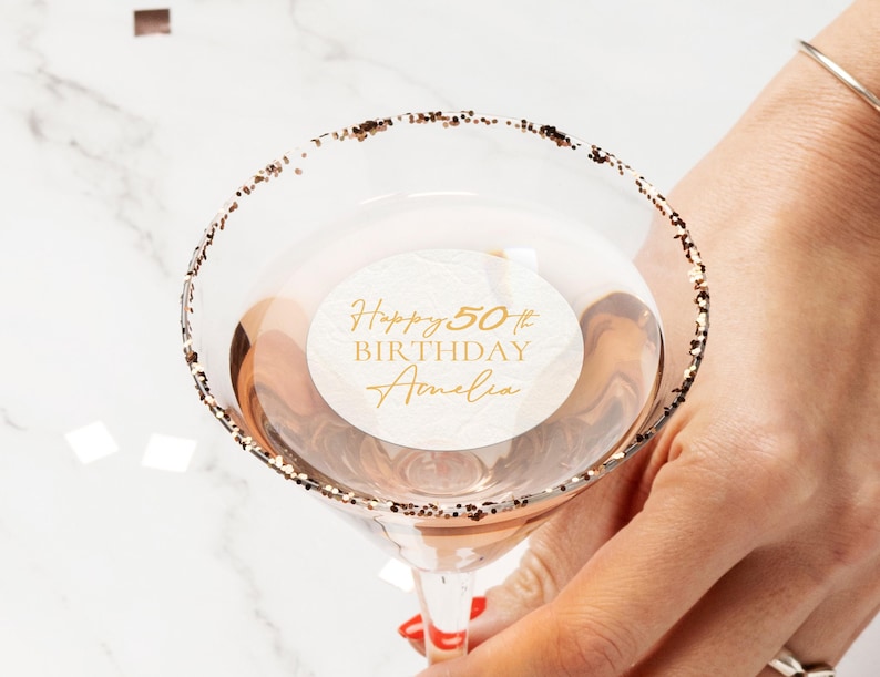 May include: A martini glass with a gold glitter rim, filled with a light-coloured liquid. A white circular sticker inside the glass reads "Happy 50th Birthday Amelia" in gold script. The glass is held by a person.