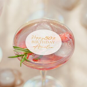 May include: A cocktail in a stemmed glass with ice, a sprig of rosemary, and a white circular label with gold text that reads "Happy 50th BIRTHDAY Amelia". The drink is pink and appears to have fruit in it.