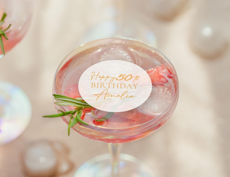 May include: A cocktail in a stemmed glass with ice, a sprig of rosemary, and a white circular label with gold text that reads "Happy 50th BIRTHDAY Amelia". The drink is pink and appears to have fruit in it.