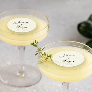 May include: Two coupe glasses filled with a pale yellow cocktail. Each glass has a white circular sticker with the names "Jason Liya" and the date "07.12.25". A sprig of green herbs adorns one glass. The background is a soft gray.
