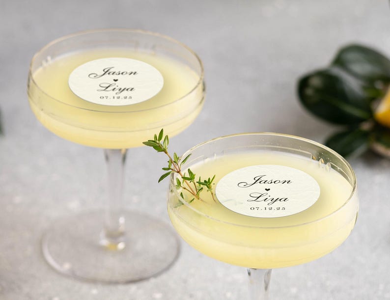 May include: Two coupe glasses filled with a pale yellow cocktail. Each glass has a white circular sticker with the names "Jason Liya" and the date "07.12.25". A sprig of green herbs adorns one glass. The background is a soft gray.