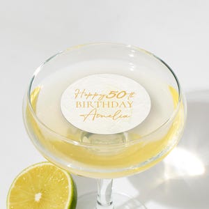 May include: A champagne glass with a yellow cocktail and a white label inscribed with "Happy 50th Birthday Amelia" in gold. A lime wedge is placed beside the glass. The image has a celebratory theme.