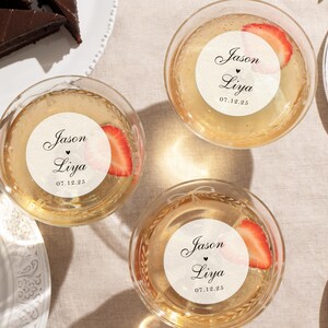 May include: Overhead shot of three round glasses filled with a golden liquid, each with a strawberry slice and a circular sticker that reads "Jason Liya 07.12.25". Slices of chocolate cake are on white plates.