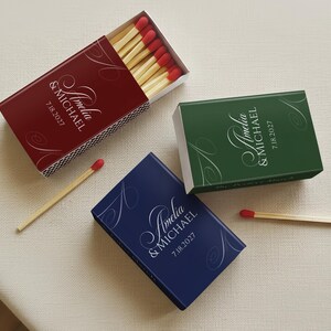 May include: Three matchboxes in red, green, and blue, each with the names "Amelia & Michael" and the date "7.18.2027" in elegant script. The red box is open, revealing matches with red tips. Decorative swirls accent the text.