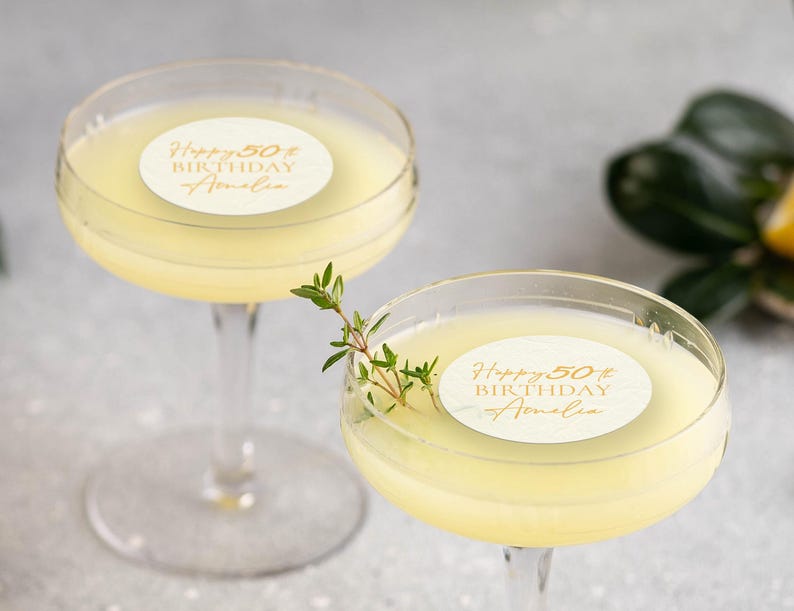 May include: Two clear coupe glasses filled with a pale yellow cocktail. Each drink has a round sticker with gold text that reads "Happy 50th Birthday Amelia." One glass is garnished with a sprig of green herbs. The background is a soft grey.